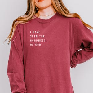 May include: A maroon long-sleeve shirt with the text "I HAVE SEEN THE GOODNESS OF GOD" in white block letters. The shirt has a crew neck and a relaxed fit. The fabric appears soft and comfortable.