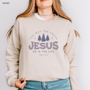 May include: Sand-colored sweatshirt with the text "THE WAY THE TRUTH JESUS HE IS THE LIFE John 14:6" in a distressed purple font. The design includes three stylized pine trees. The sweatshirt is a crew neck style.