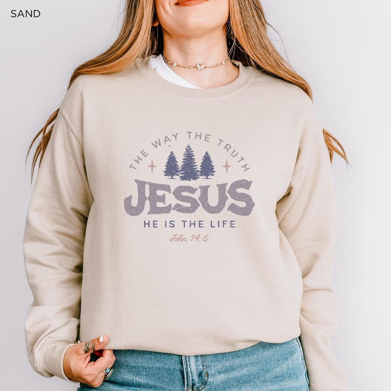 May include: Sand-colored sweatshirt with the text "THE WAY THE TRUTH JESUS HE IS THE LIFE John 14:6" in a distressed purple font. The design includes three stylized pine trees. The sweatshirt is a crew neck style.