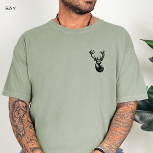 May include: A sage green t-shirt with a black deer graphic on the front. The deer has large antlers and is looking to the right.