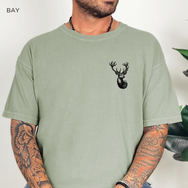 May include: A sage green t-shirt with a black deer graphic on the front. The deer has large antlers and is looking to the right.