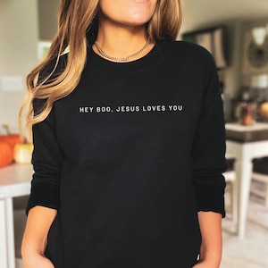 May include: Black crew neck sweatshirt with the white text "HEY BOO, JESUS LOVES YOU". The long-sleeved top is made of a soft material. The sweatshirt is a casual style.