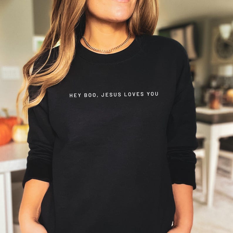 May include: Black crew neck sweatshirt with the white text "HEY BOO, JESUS LOVES YOU". The long-sleeved top is made of a soft material. The sweatshirt is a casual style.