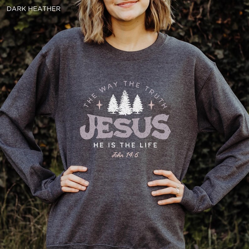 May include: Dark heather gray sweatshirt with the text "THE WAY THE TRUTH JESUS HE IS THE LIFE John 14:6" in a distressed, light purple font. The design includes three white pine trees and stars. The sweatshirt is a long-sleeved crew neck.