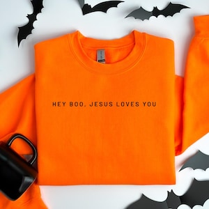 May include: Bright orange sweatshirt with the text "HEY BOO, JESUS LOVES YOU" in black. The sweatshirt is surrounded by black bat decorations and a black mug.