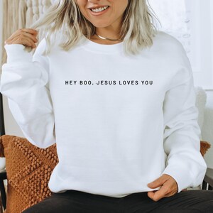 May include: White sweatshirt with the text "HEY BOO, JESUS LOVES YOU" in black. The long-sleeved top is made of a soft material and is suitable for casual wear. The sweatshirt is a comfortable and stylish choice for everyday wear.