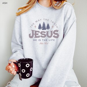 May include: Ash gray sweatshirt with the text "THE WAY THE TRUTH JESUS HE IS THE LIFE John 14:6" in a circular design. The sweatshirt features a graphic of three trees. A black mug with white flower designs is held in the hand.