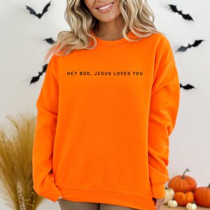 May include: Bright orange sweatshirt with the text "HEY BOO, JESUS LOVES YOU" in black. The sweatshirt is a solid color and has a crew neck. The background includes Halloween decorations such as bats and pumpkins.