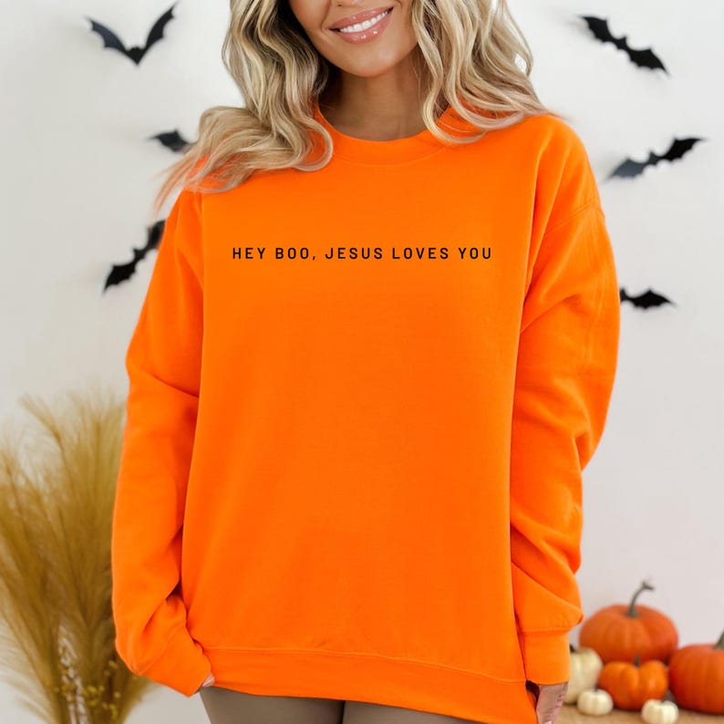 May include: Bright orange sweatshirt with the text "HEY BOO, JESUS LOVES YOU" in black. The sweatshirt is a solid color and has a crew neck. The background includes Halloween decorations such as bats and pumpkins.