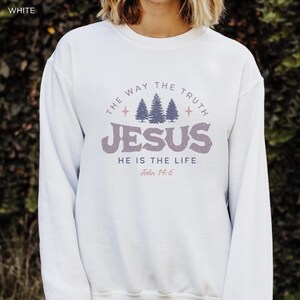 May include: White sweatshirt with the text "THE WAY THE TRUTH JESUS HE IS THE LIFE John 14:6" in a circular design. The design includes three stylized pine trees and star accents, in shades of purple and gray.