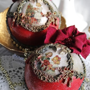 Puede incluir: Dos adornos navideños esféricos con una base roja de acabado brillante y una sección superior de color crema. Cada adorno está decorado con una ilustración de estilo vintage de niños, un borde texturizado dorado y un lazo de tela rojo oscuro.