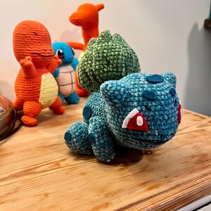 Op de afbeelding: Een verzameling handgemaakte gehaakte Pokémon-figuren. De voorgrond toont een teal Bulbasaur met rode en witte accenten. Achter hem staan oranje Charmander, blauwe Squirtle en oranje Charizard haakfiguren. De figuren worden op een houten oppervlak gepresenteerd.