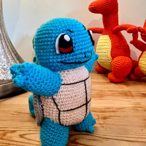 May include: A handmade crochet Squirtle plush toy in shades of blue, with a beige shell and black accents. The toy has large, expressive eyes and is posed with its arms outstretched. Other orange and yellow crochet figures are in the background.