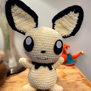 May include: A handmade, cream-colored plush bunny with large black-trimmed ears and big eyes. The bunny has a black bow tie and pink cheeks. It is sitting on a wooden surface.
