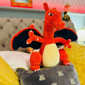 Könnte beinhalten: Ein großer, handgefertigter orangefarbener Häkeldrache mit schwarzen und blauen Flügeln und einem cremefarbenen Bauch. Der Drache sitzt auf einem Bett mit gelben und grauen Kissen. Die Krallen und Hörner des Drachen sind weiß.