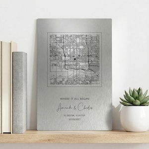 May include: A silver metal print featuring a black and white city map. The text "WHERE IT ALL BEGAN" is above the names "Amanda & Charles", with coordinates and a date below. A succulent in a white pot is on a wooden shelf.