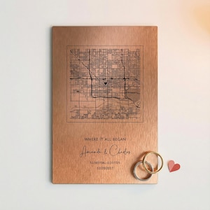 May include: A copper-colored metal plaque with a detailed map engraving. The text "WHERE IT ALL BEGAN" is above the names "Amanda & Charles". Two rings and a small heart are next to the plaque. The date 12/20/2017 is also included.