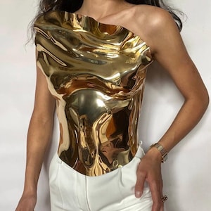 Puede incluir: Un top dorado met&aacute;lico de un solo hombro con textura ondulada. Se combina con pantalones blancos. La superficie reflectante crea un efecto l&iacute;quido. El estilo es moderno y vanguardista, ideal para un look audaz.
