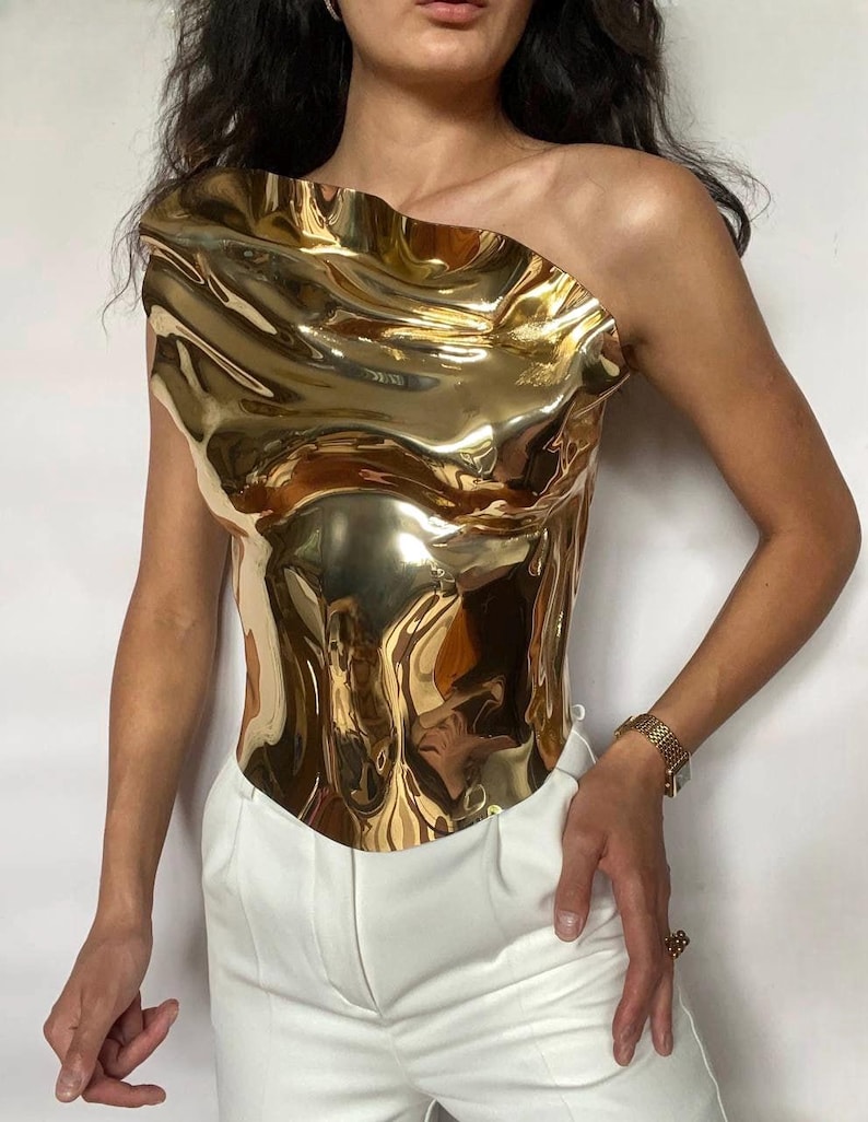 Puede incluir: Un top dorado met&aacute;lico de un solo hombro con textura ondulada. Se combina con pantalones blancos. La superficie reflectante crea un efecto l&iacute;quido. El estilo es moderno y vanguardista, ideal para un look audaz.