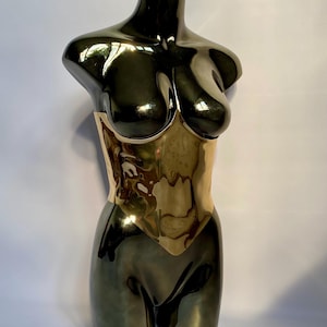 Puede incluir: Un maniqu&iacute; de torso de metal oscuro brillante con una secci&oacute;n de cintura dorada reflectante. La escultura muestra una forma suave y contorneada con un acabado de alto brillo que captura la luz en su superficie curva, resaltando las &aacute;reas del pecho y las caderas.