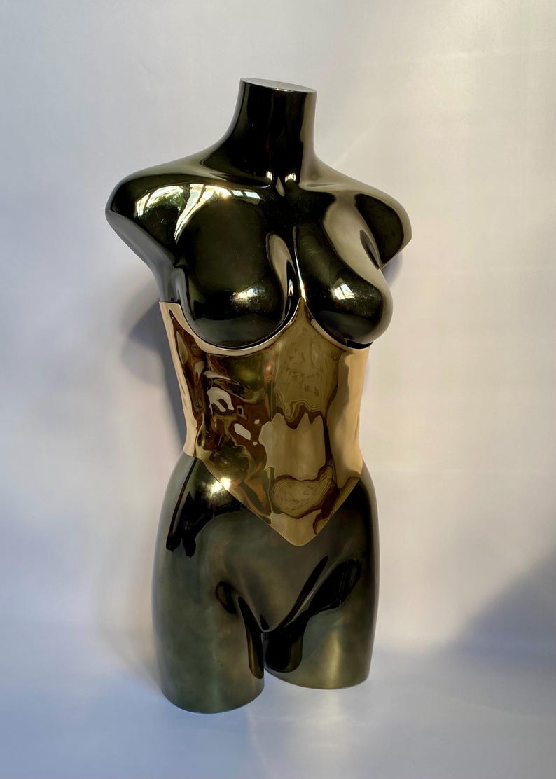Puede incluir: Un maniqu&iacute; de torso de metal oscuro brillante con una secci&oacute;n de cintura dorada reflectante. La escultura muestra una forma suave y contorneada con un acabado de alto brillo que captura la luz en su superficie curva, resaltando las &aacute;reas del pecho y las caderas.