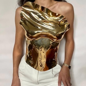 Puede incluir: Un top dorado met&aacute;lico de un solo hombro con un dise&ntilde;o escultural ondulado. El top se combina con pantalones blancos. La superficie reflectante del top crea un efecto visual din&aacute;mico, destacando su forma y textura &uacute;nicas. Se ve un reloj dorado en la mu&ntilde;eca.