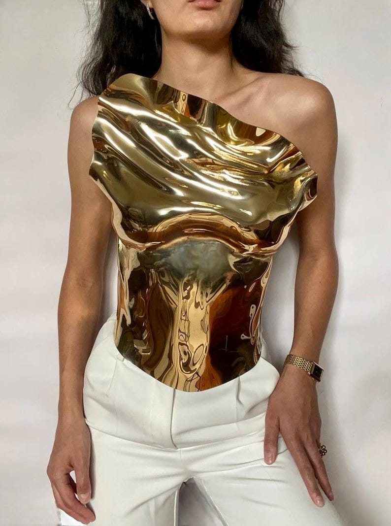 Puede incluir: Un top dorado met&aacute;lico de un solo hombro con un dise&ntilde;o escultural ondulado. El top se combina con pantalones blancos. La superficie reflectante del top crea un efecto visual din&aacute;mico, destacando su forma y textura &uacute;nicas. Se ve un reloj dorado en la mu&ntilde;eca.