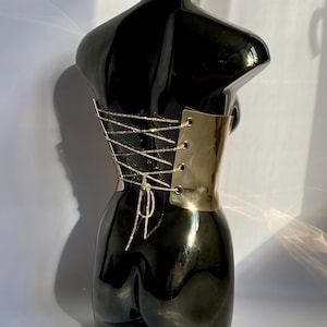 May include: A black, glossy torso sculpture with a gold-colored metal corset. The corset features a crisscross design with sparkling silver-colored string. The sculpture is displayed against a white background.