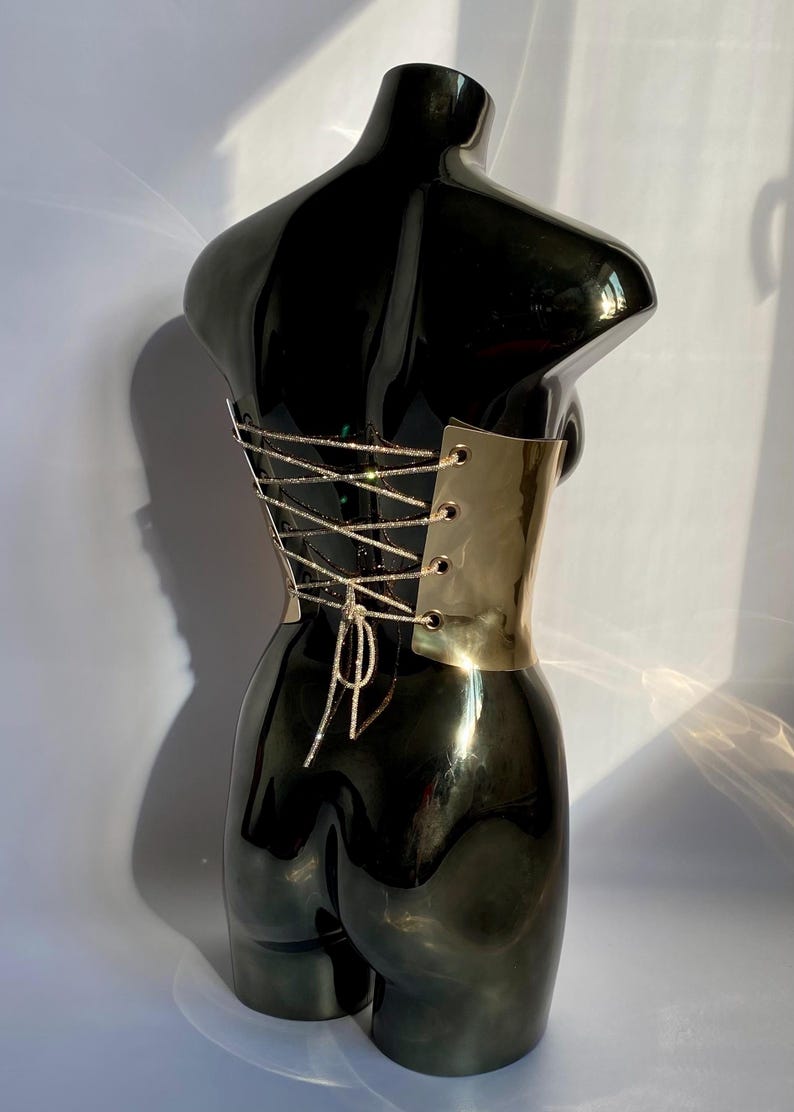 May include: A black, glossy torso sculpture with a gold-colored metal corset. The corset features a crisscross design with sparkling silver-colored string. The sculpture is displayed against a white background.