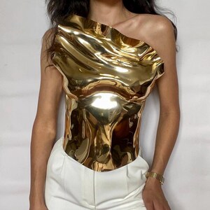 Puede incluir: Un top dorado met&aacute;lico de un solo hombro con un dise&ntilde;o escultural y ondulado. El top tiene un corpi&ntilde;o ajustado y un escote asim&eacute;trico &uacute;nico. La prenda se combina con pantalones blancos. La superficie reflectante del top crea un efecto visual llamativo.