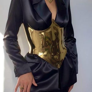 May include: A polished gold-colored corset worn over a black satin blazer and pants. The corset has a sculpted, form-fitting design. A gold necklace and ring complete the look. The outfit is a bold fashion statement.
