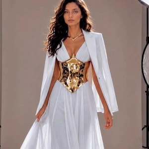 Puede incluir: Una mujer con un vestido blanco con un corsé dorado y una blazer blanca. El vestido tiene una falda fluida y un escote en V. El corsé dorado tiene un diseño intrincado. La blazer está drapeada sobre los hombros.