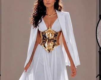 Puede incluir: Una mujer con un vestido blanco con un corsé dorado y una blazer blanca. El vestido tiene una falda fluida y un escote en V. El corsé dorado tiene un diseño intrincado. La blazer está drapeada sobre los hombros.