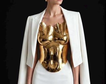 Peut inclure: Une femme porte une robe et un blazer blancs, avec un bustier doré sculpté. Le bustier présente un design organique détaillé. Le fond est noir, soulignant le contraste de la tenue.