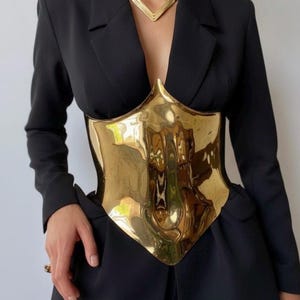 May include: A gold-colored metal corset-style top with a reflective surface. The corset is worn over a black blazer and paired with a matching gold necklace. The overall look is a bold fashion statement.