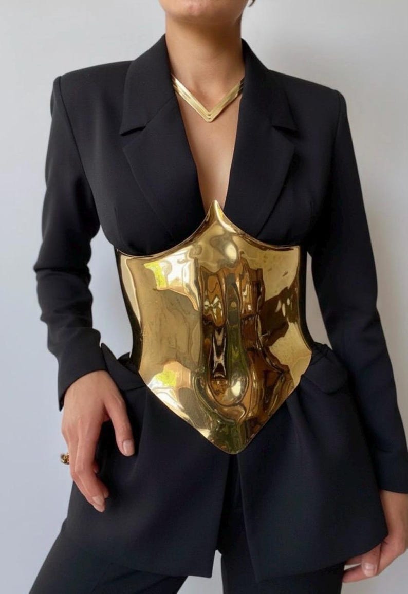 May include: A gold-colored metal corset-style top with a reflective surface. The corset is worn over a black blazer and paired with a matching gold necklace. The overall look is a bold fashion statement.