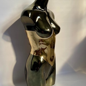 Puede incluir: Un maniqu&iacute; de torso de metal oscuro y brillante con una pieza de cors&eacute; de color dorado altamente reflectante en la cintura. El maniqu&iacute; tiene una forma suave y contorneada con un busto y una cintura definidos, con un acabado pulido.