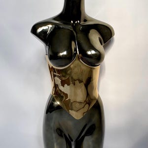 May include: A metallic sculpture of a female torso. The upper portion is a dark, reflective color, transitioning to a gold-toned midsection. The sculpture is smooth and polished, with a minimalist design.