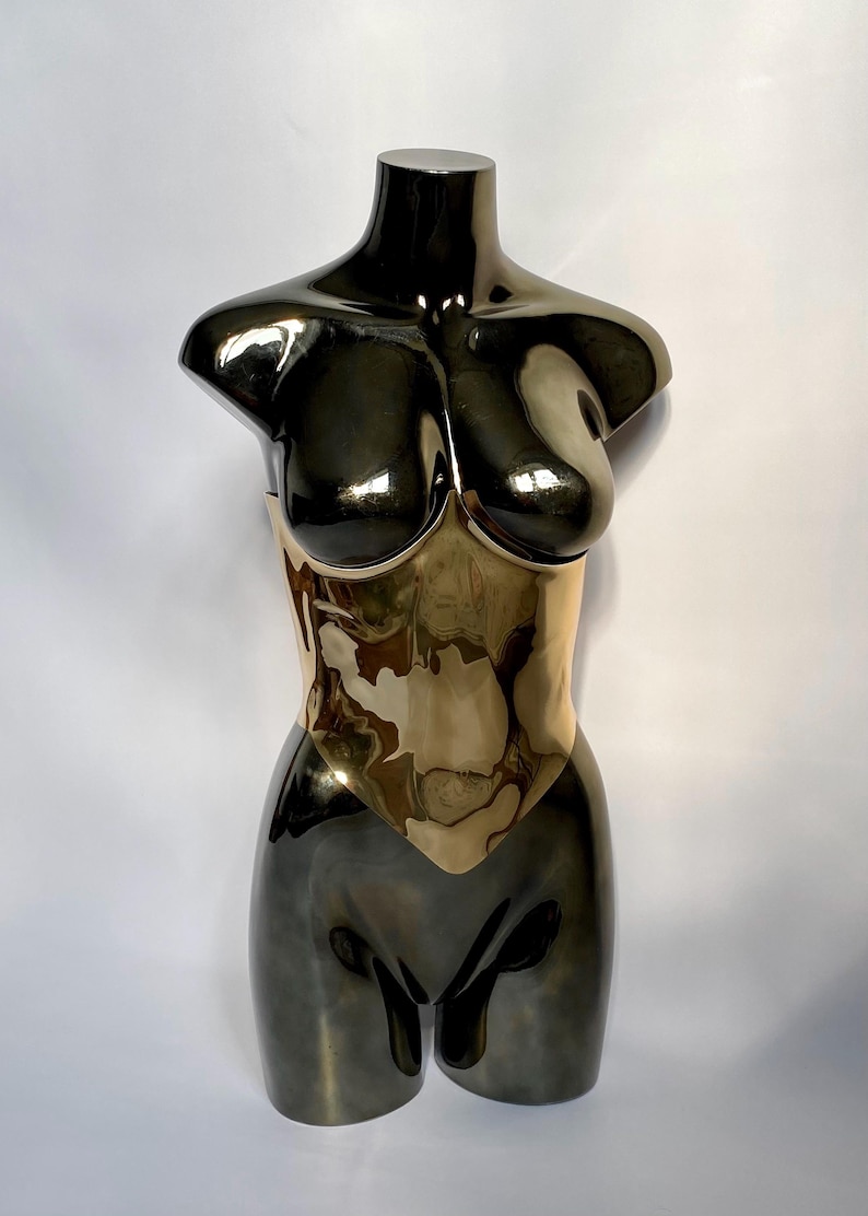May include: A metallic sculpture of a female torso. The upper portion is a dark, reflective color, transitioning to a gold-toned midsection. The sculpture is smooth and polished, with a minimalist design.