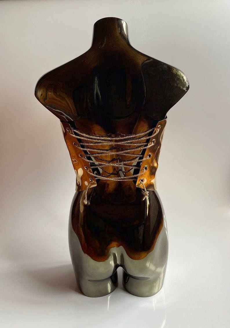 Puede incluir: Escultura decorativa de torso con acabado marr&oacute;n oscuro brillante. La escultura presenta un dise&ntilde;o tipo cors&eacute; en la parte posterior con cordones plateados. La parte inferior de la escultura tiene un acabado gris met&aacute;lico.