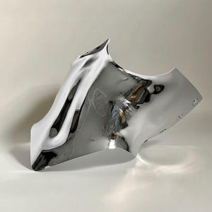 Puede incluir: Una escultura abstracta moderna con un acabado plateado metálico reflectante. La pieza tiene una forma fluida y orgánica con bordes curvos y una superficie ondulada, creando un efecto visual dinámico. La escultura se exhibe sobre una superficie blanca.
