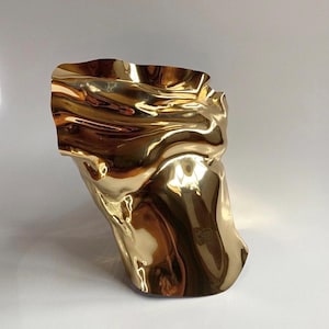 Puede incluir: Una escultura abstracta dorada con un dise&ntilde;o ondulado y fluido. La superficie met&aacute;lica refleja la luz, creando reflejos y sombras. La forma es org&aacute;nica y parece ser una representaci&oacute;n estilizada de un torso.