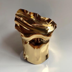 Puede incluir: Una escultura abstracta de color dorado con una parte superior ondulada en forma de ola. El acabado met&aacute;lico refleja la luz, creando reflejos y sombras. La base tiene forma cil&iacute;ndrica y el dise&ntilde;o general es moderno y art&iacute;stico.