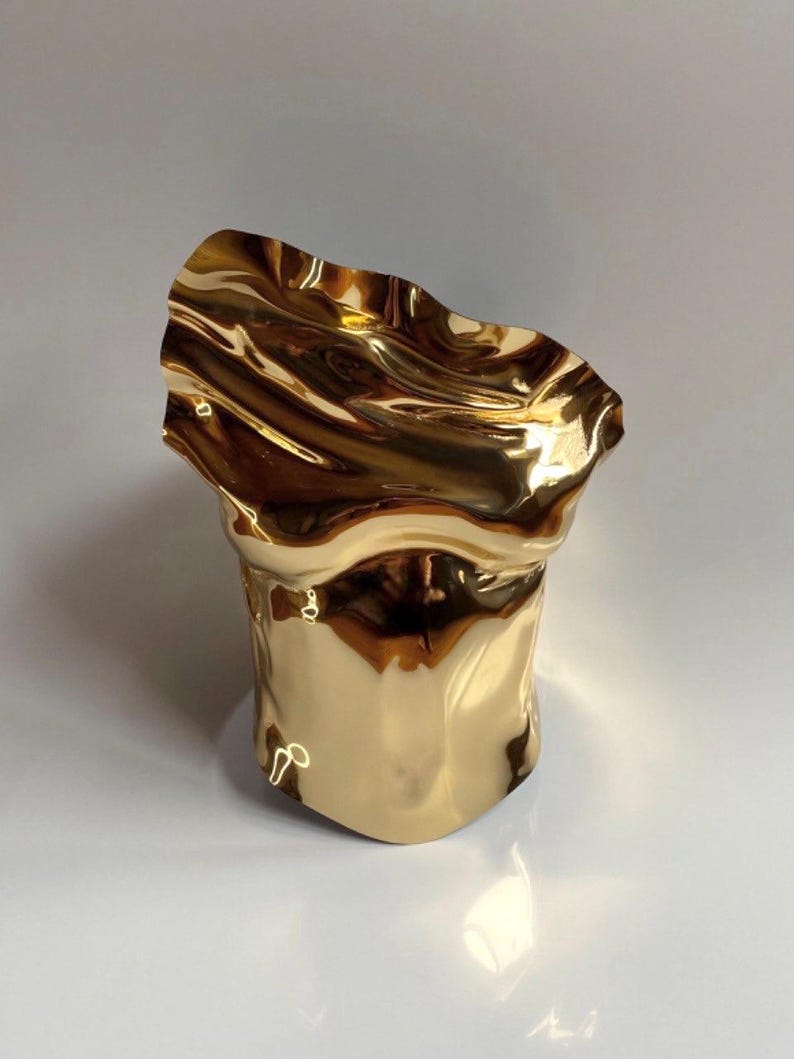 Puede incluir: Una escultura abstracta de color dorado con una parte superior ondulada en forma de ola. El acabado met&aacute;lico refleja la luz, creando reflejos y sombras. La base tiene forma cil&iacute;ndrica y el dise&ntilde;o general es moderno y art&iacute;stico.