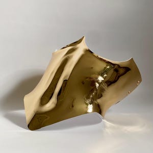 May include: A gold-colored, abstract sculpture with a reflective, rippled surface. The piece has a curved shape with a pointed top and a smooth, polished finish. The sculpture is set against a plain white background.