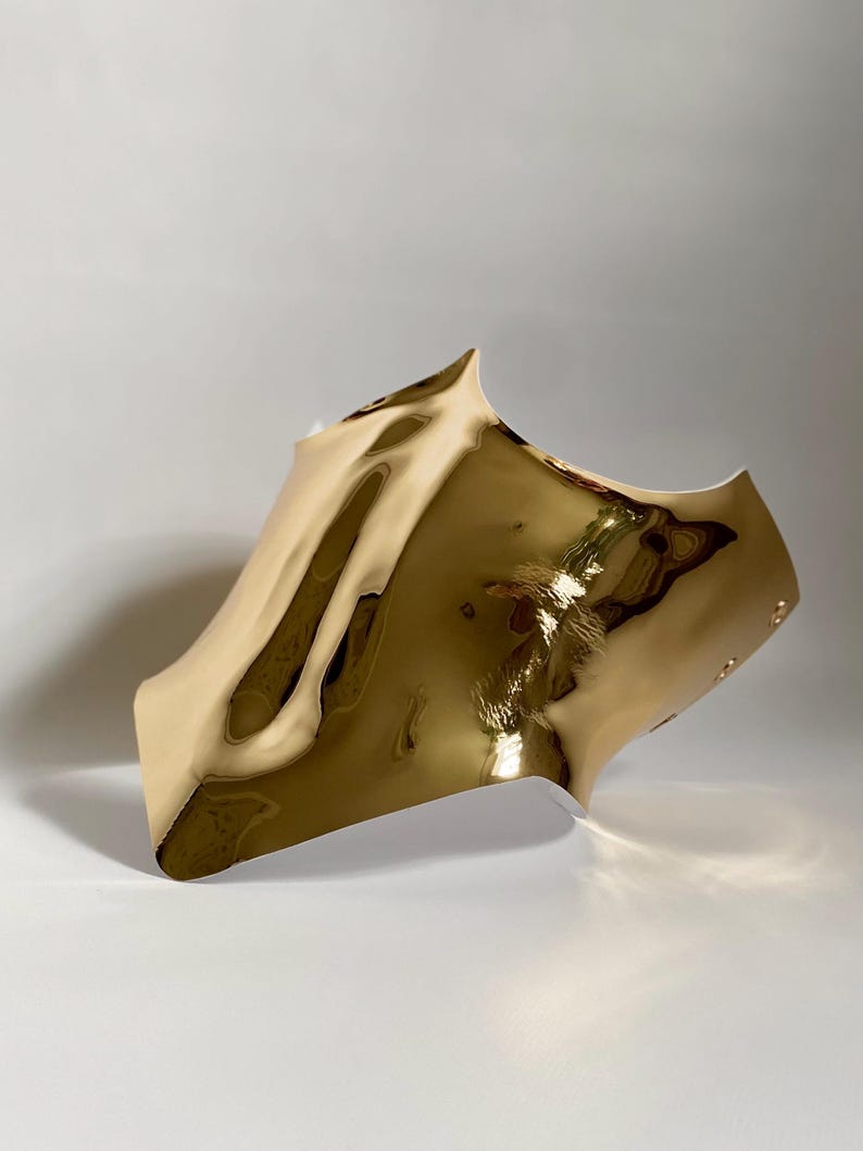 May include: A gold-colored, abstract sculpture with a reflective, rippled surface. The piece has a curved shape with a pointed top and a smooth, polished finish. The sculpture is set against a plain white background.