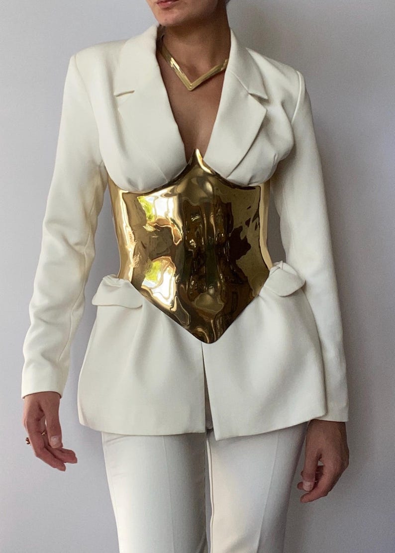 Puede incluir: Un blazer color crema y pantalones a juego combinados con un cors&eacute; de metal dorado. El blazer tiene un dise&ntilde;o cl&aacute;sico con solapas de muesca. Un collar dorado completa el look. El atuendo es una pieza llamativa.