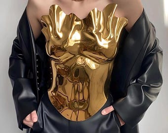 May include: A gold-colored, metallic bustier with a sculpted, organic design. The bustier is worn with black leather pants and a black leather jacket. A silver necklace is also worn.