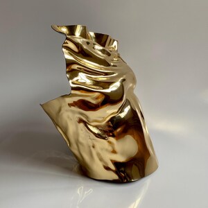 Puede incluir: Una escultura abstracta dorada con una forma fluida y drapeada. La superficie met&aacute;lica refleja la luz, creando reflejos y sombras. La escultura tiene una forma &uacute;nica y org&aacute;nica, que sugiere movimiento y fluidez. La obra de arte est&aacute; sobre un fondo neutro.
