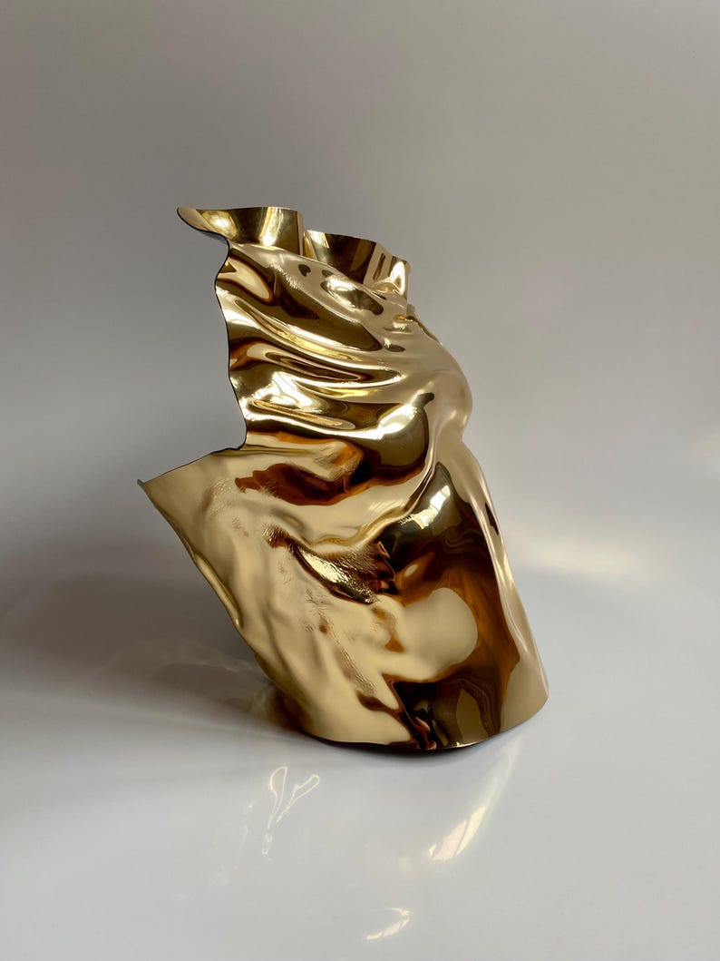 Puede incluir: Una escultura abstracta dorada con una forma fluida y drapeada. La superficie met&aacute;lica refleja la luz, creando reflejos y sombras. La escultura tiene una forma &uacute;nica y org&aacute;nica, que sugiere movimiento y fluidez. La obra de arte est&aacute; sobre un fondo neutro.